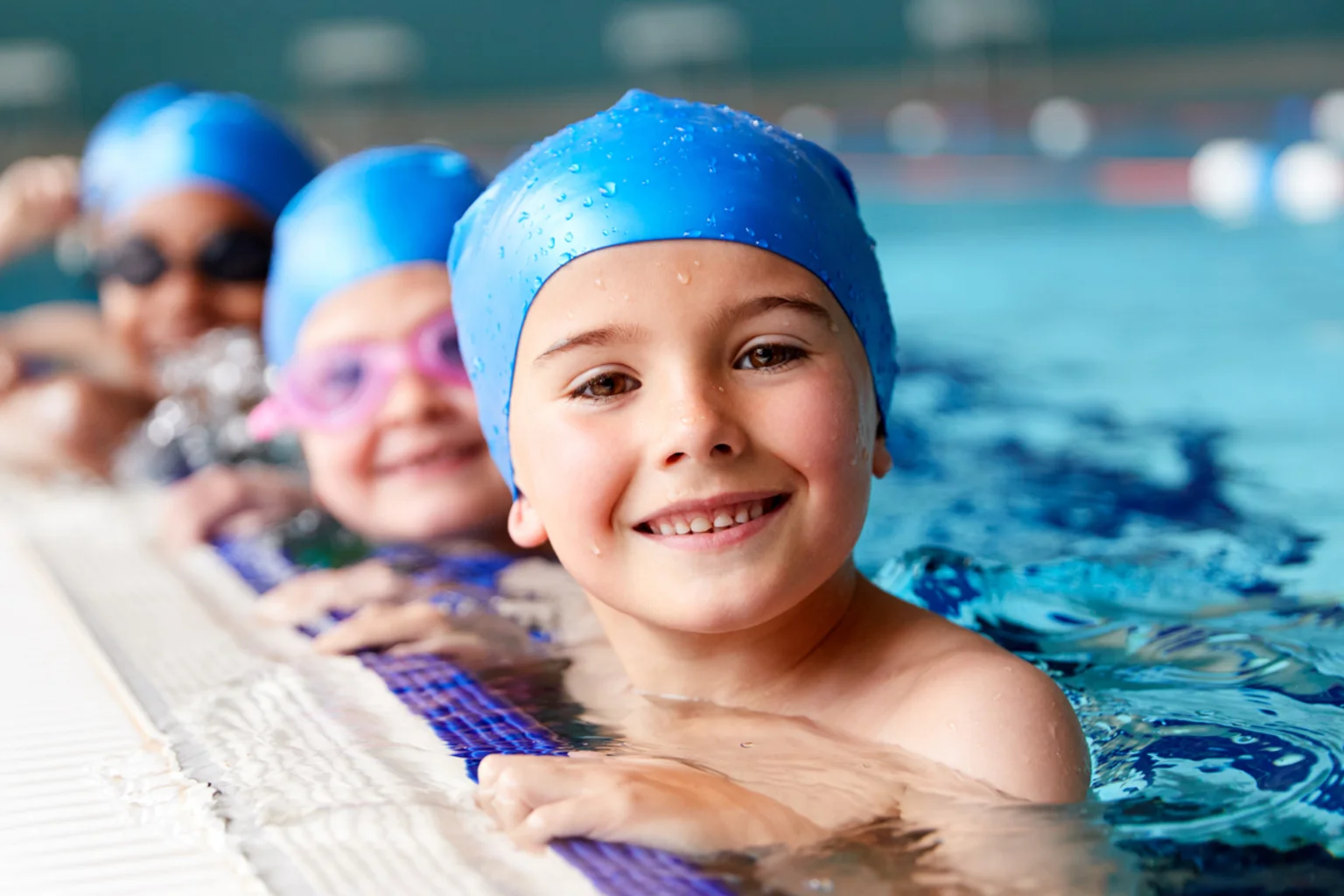 Kinder mit Badekappen lachen am Beckenrand beim Schwimmkurs